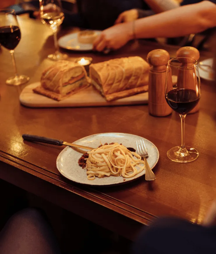 Plate of pasta with red sauce on a wooden table next to a glass of red wine, bread loaves, and salt and pepper shakers.