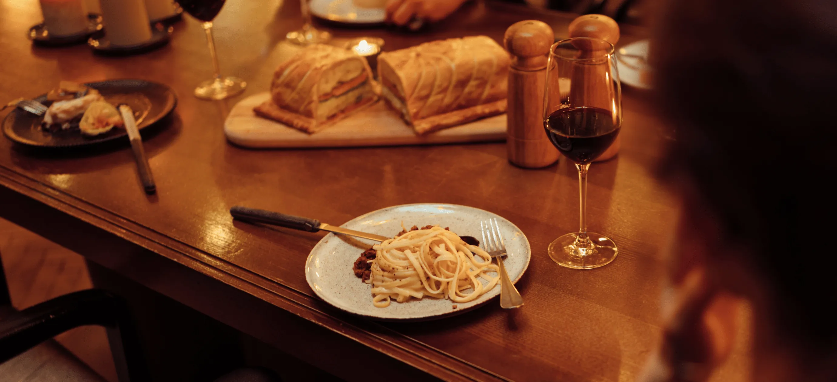 Table setting with a plate of pasta, a glass of red wine, sliced bread on a wooden board, and salt and pepper shakers.