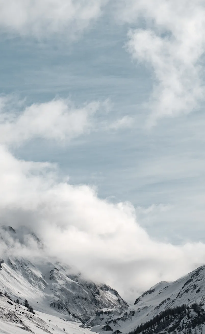 Snow-covered mountain peaks partially obscured by low clouds under a blue sky.