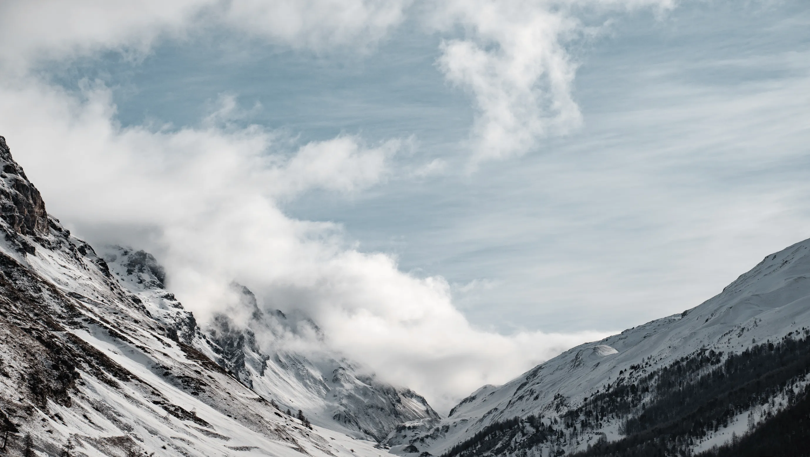 Snow-covered mountain peaks partially obscured by clouds under a cloudy blue sky.