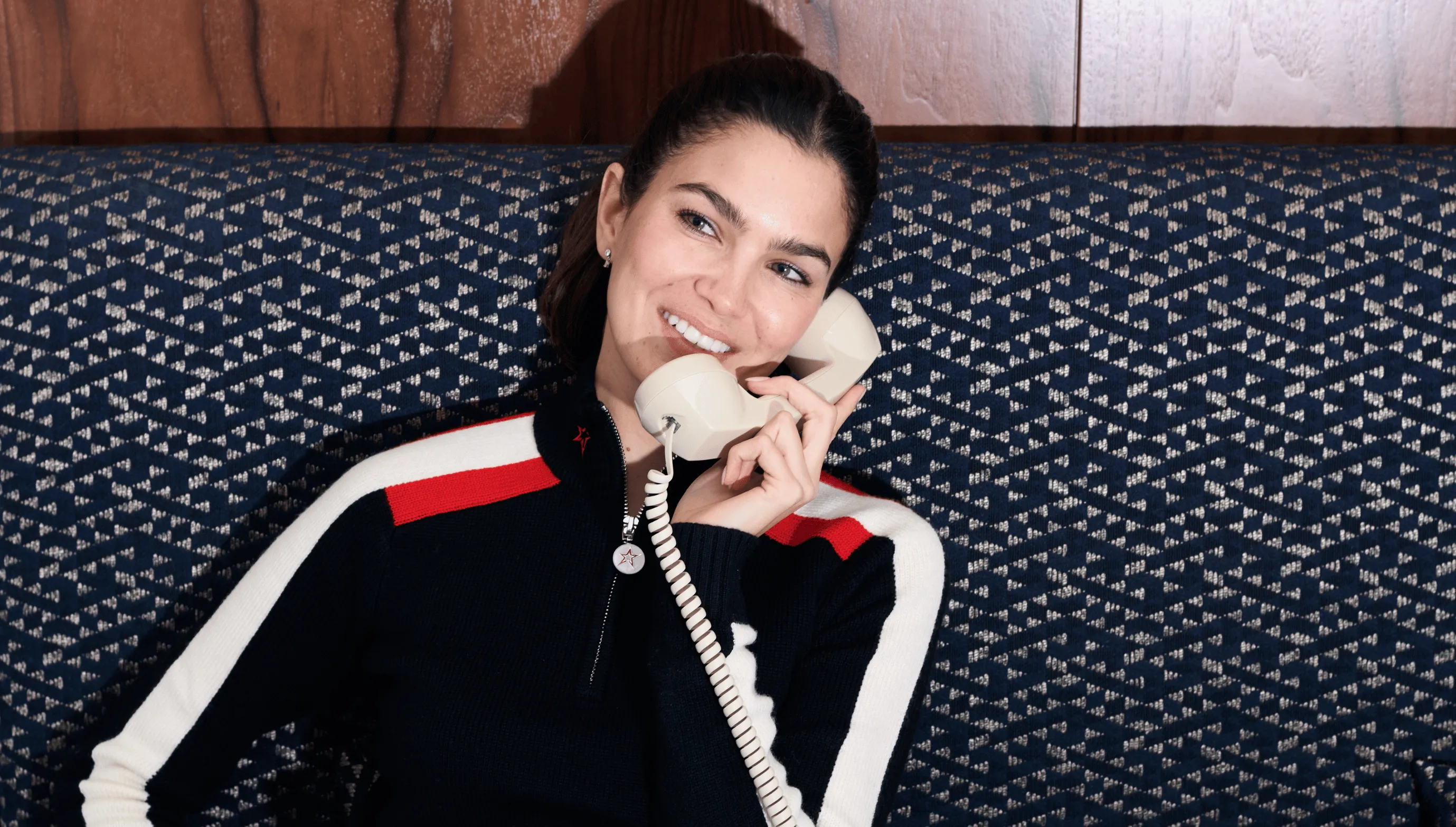 Smiling woman with dark hair wearing a black sweater with white and red stripes talking on a cream-colored corded telephone while sitting on a patterned blue sofa.