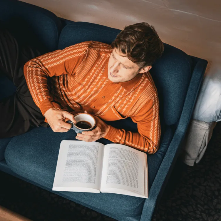 Man in an orange patterned sweater lying on a blue sofa holding a cup of coffee with an open book in front of him.