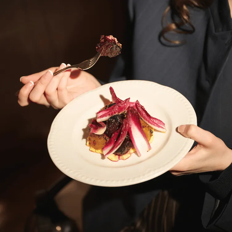 Person holding a white plate with a gourmet dish topped with red radicchio leaves and a fork with a bite of food.