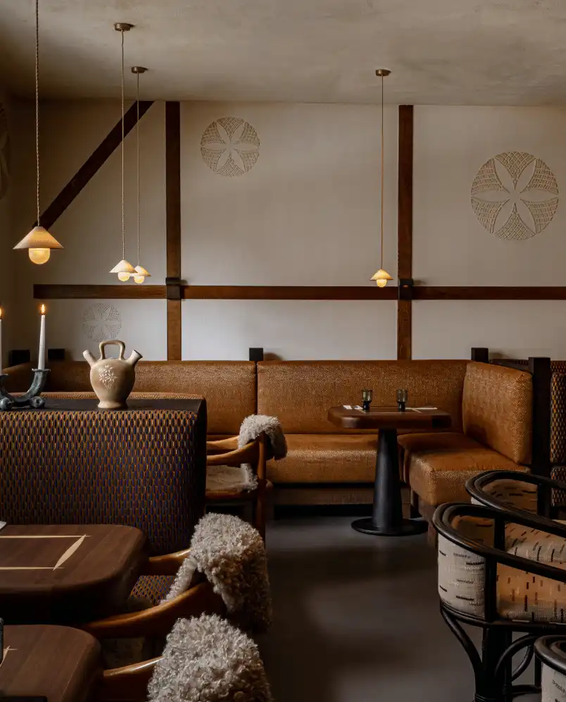 Cozy restaurant interior with brown cushioned benches, wooden tables, chairs with fur throws, hanging pendant lights, and decorative wall accents.