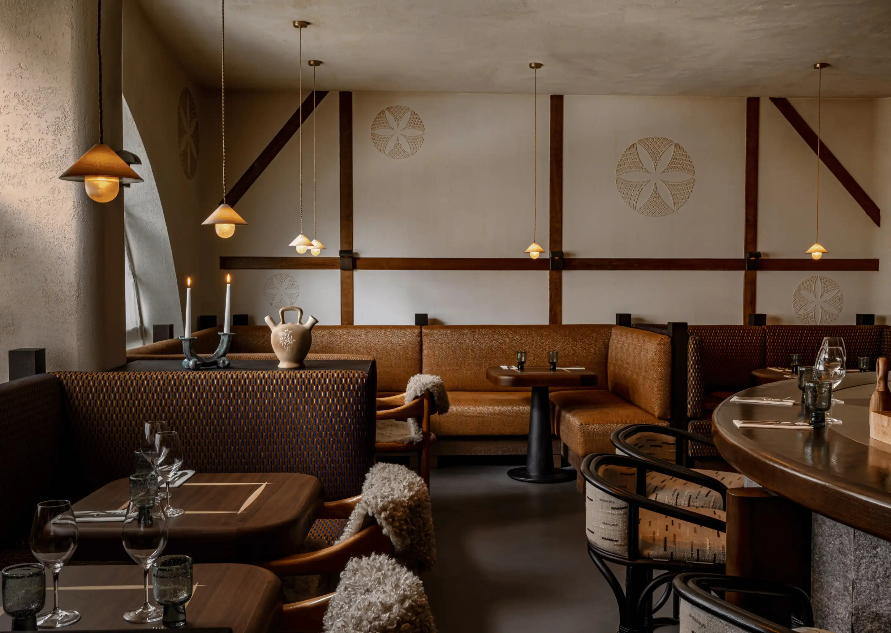 Cozy restaurant interior with brown cushioned seating, wooden tables with glassware, pendant lights, and soft candlelight.