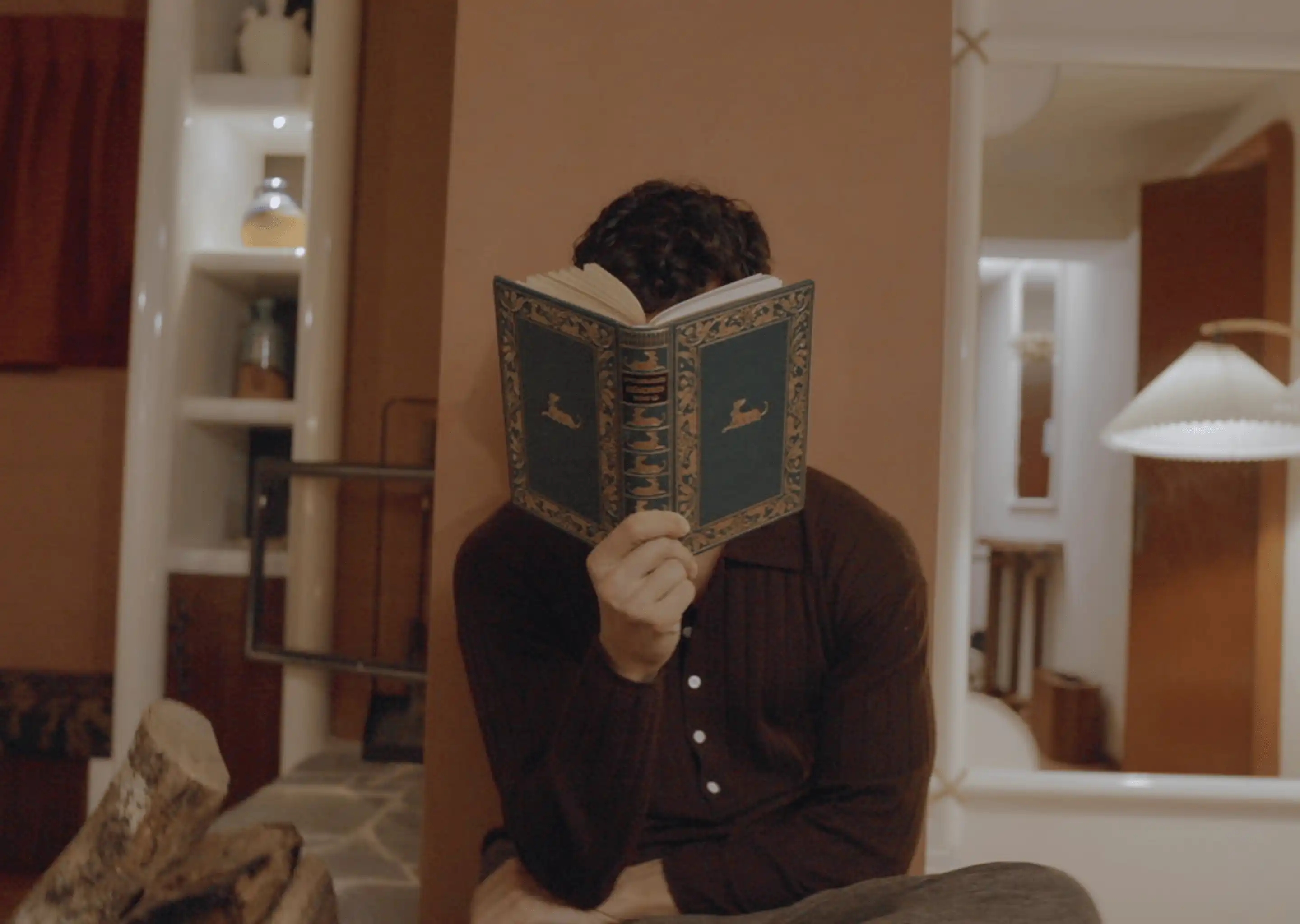 Person wearing a dark shirt sitting indoors reading an old ornate book that covers their face.