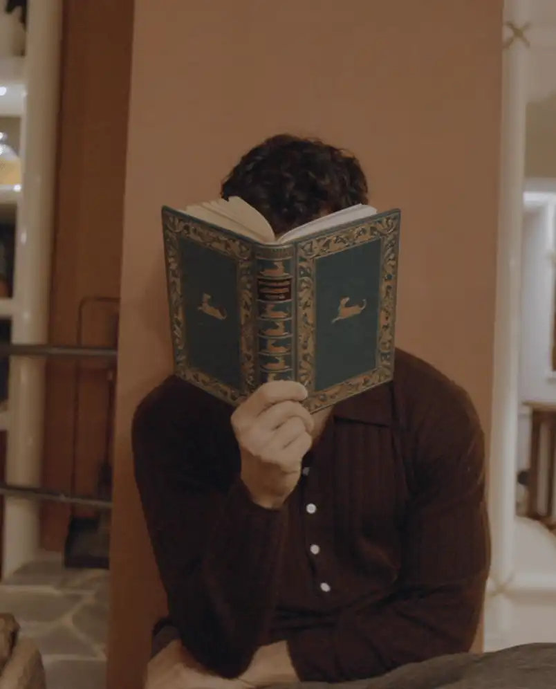 Person with curly hair sitting and reading a green hardcover book with decorative patterns and gold accents, covering their face.