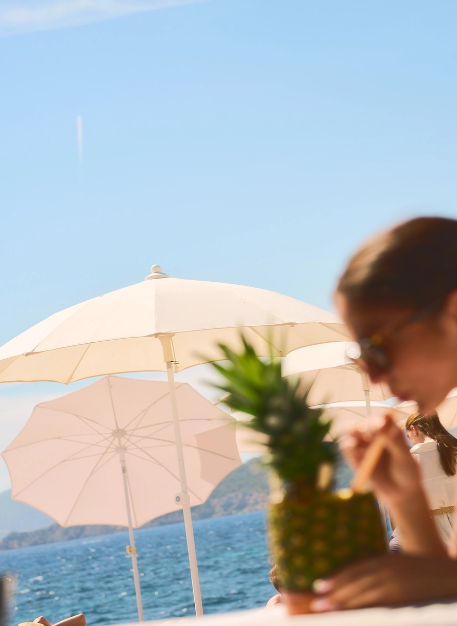 Escena de playa con sombrillas blancas, mar azul y una persona bebiendo una bebida de una taza de piña.