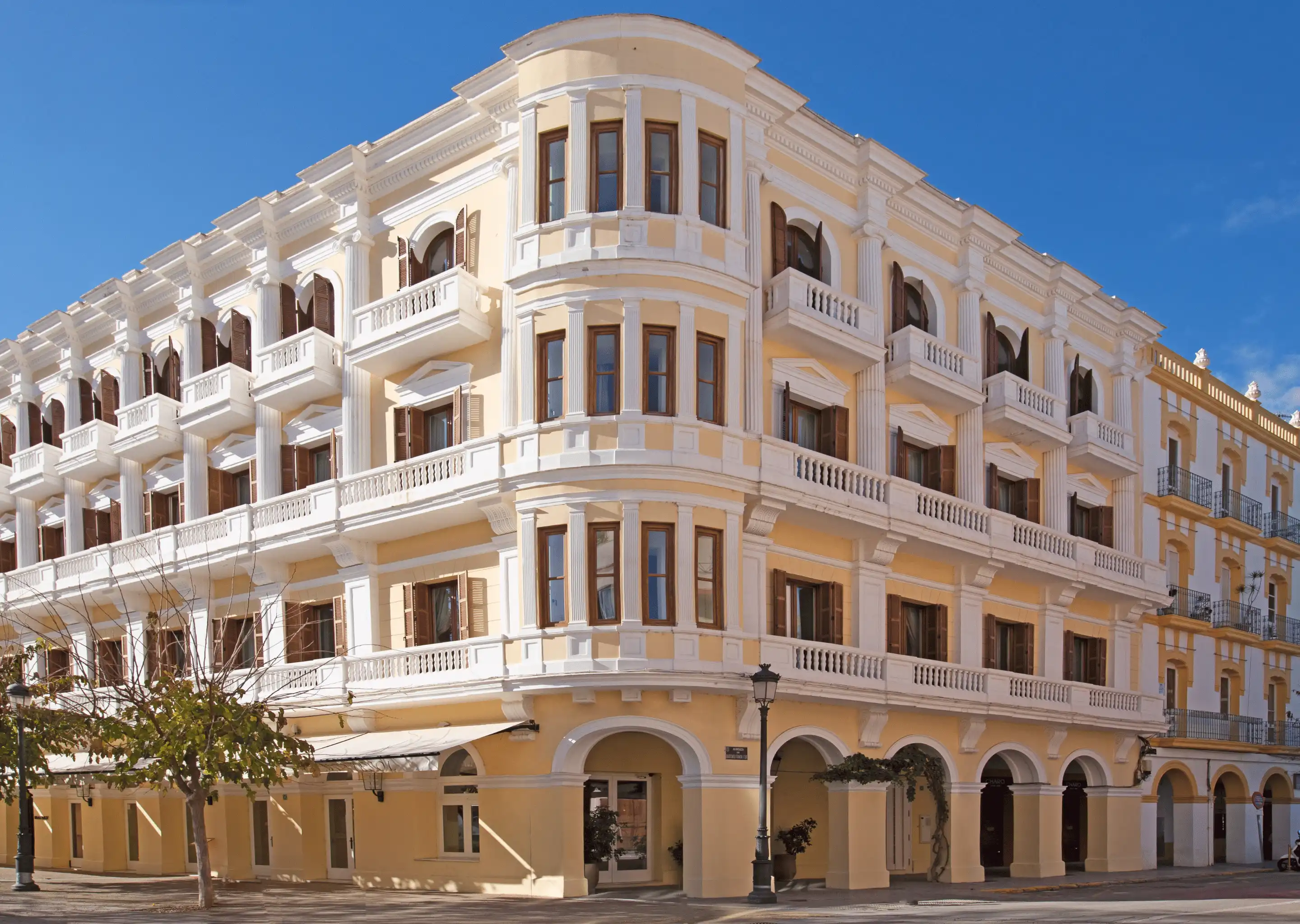 Elegant yellow and white three-story hotel building with balconies and arched doorways on a clear sunny day.