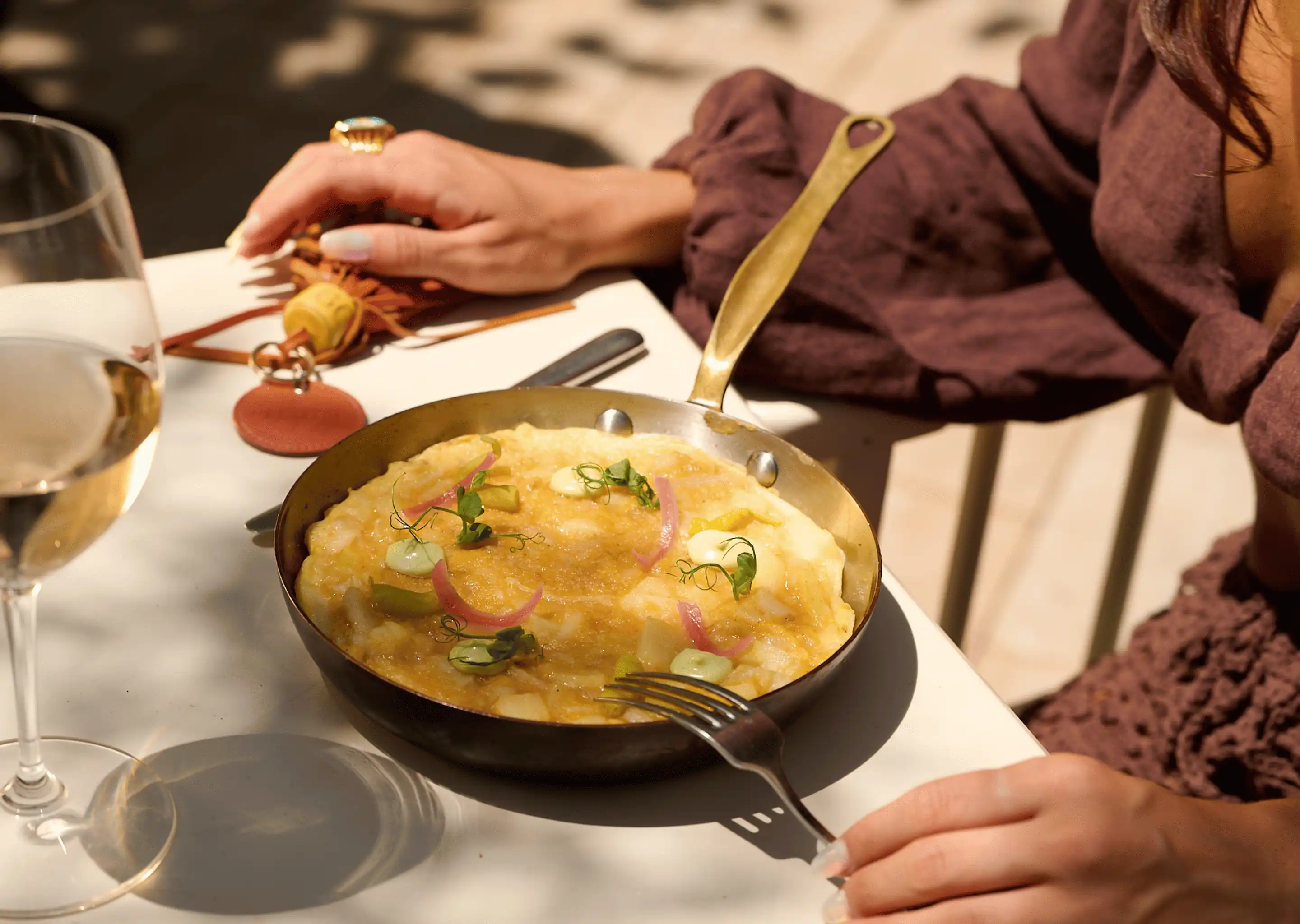 Person holding a fork near a skillet with a gourmet omelette garnished with herbs and pickled onions on a white table next to a glass of white wine.