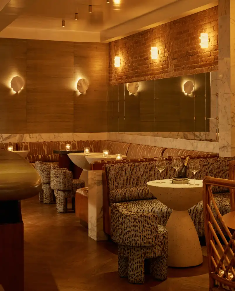 Cozy, dimly lit hotel bar seating area with patterned chairs, small tables with candles, and a bottle of wine with glasses on a table.