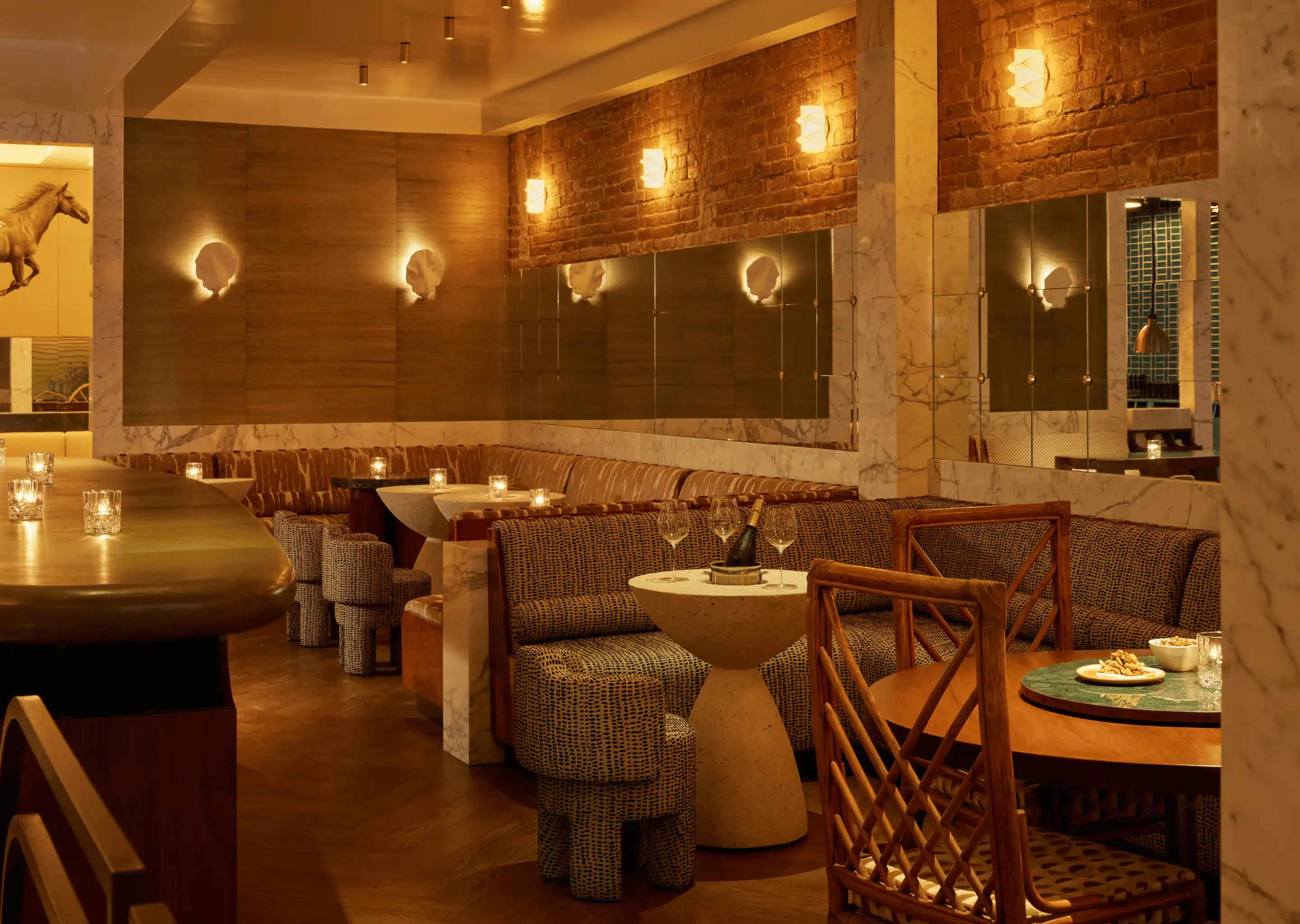 Cozy, dimly lit bar and lounge area with patterned sofas, marble tables, warm wall lights, and a bottle of champagne on a small table.
