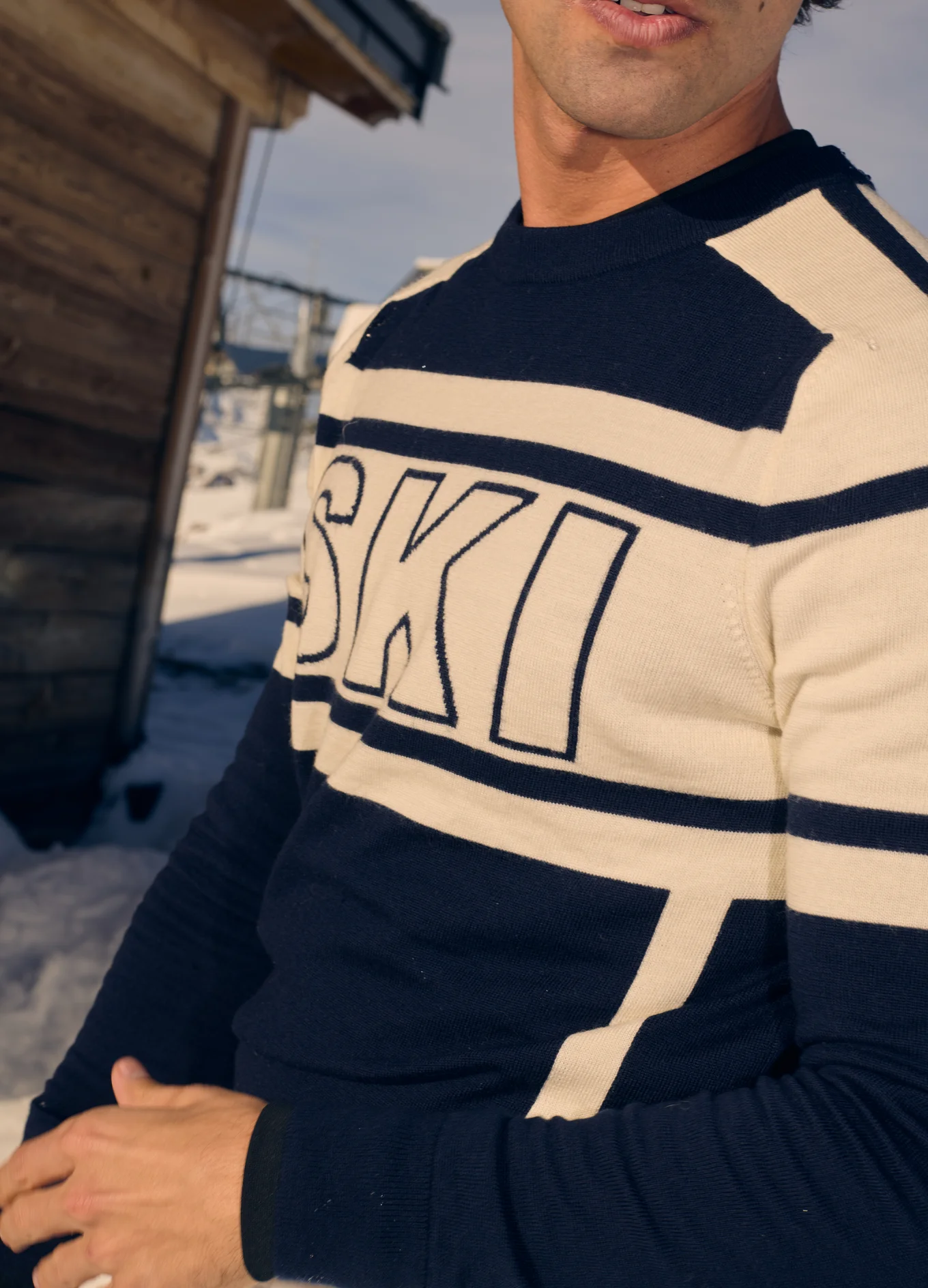 Close-up of a person wearing a navy and cream striped sweater with the word 'SKI' on it, standing outdoors with snow and a wooden cabin in the background.