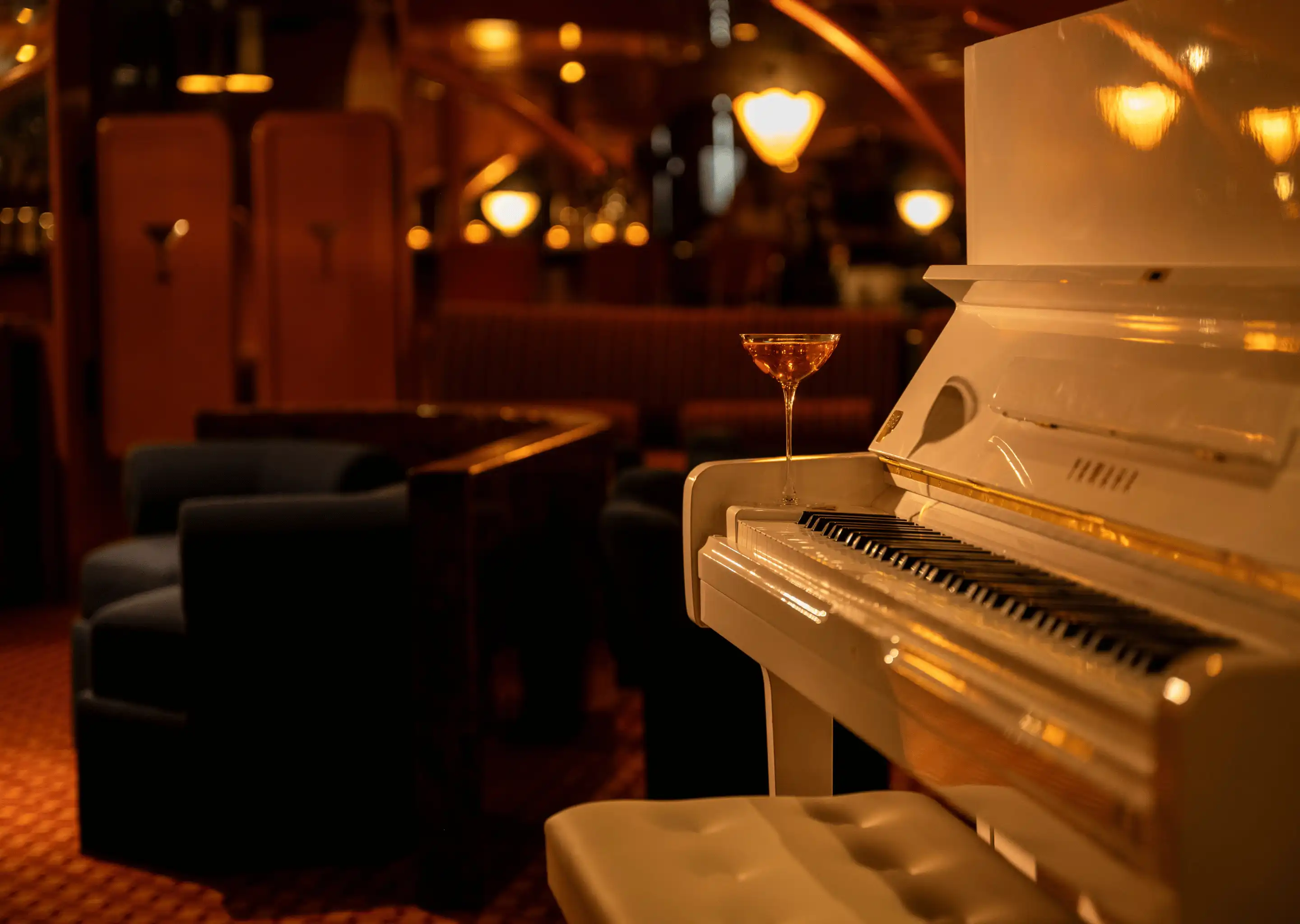 White piano with a cocktail glass placed on top in a dimly lit lounge with dark chairs and warm lighting.