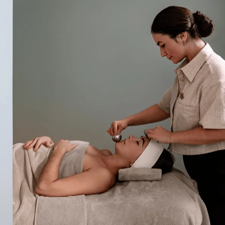 A woman receiving a facial massage with a metal tool from a spa therapist in a wellness setting.