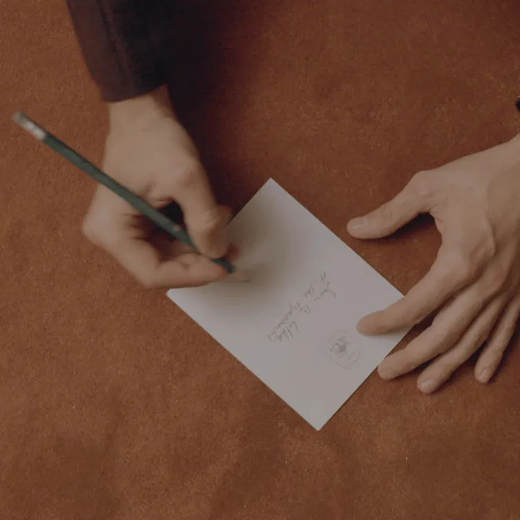 A person writing on a small white card with a pencil on a brown surface.