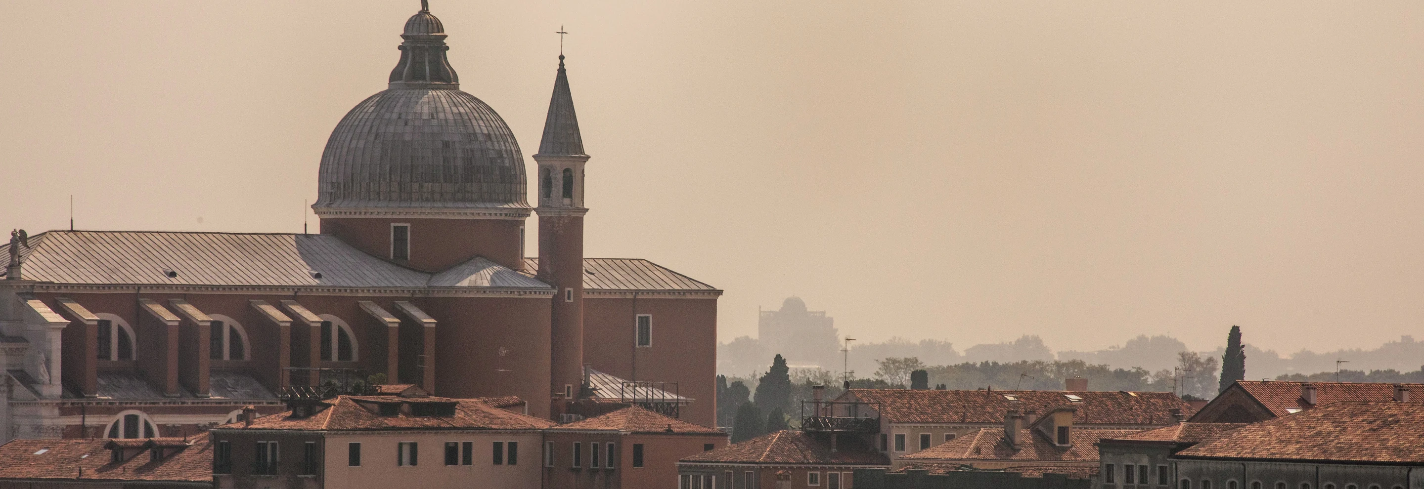 Venetian skyline featuring the dome and bell tower of a historic church against a hazy sky at sunset.