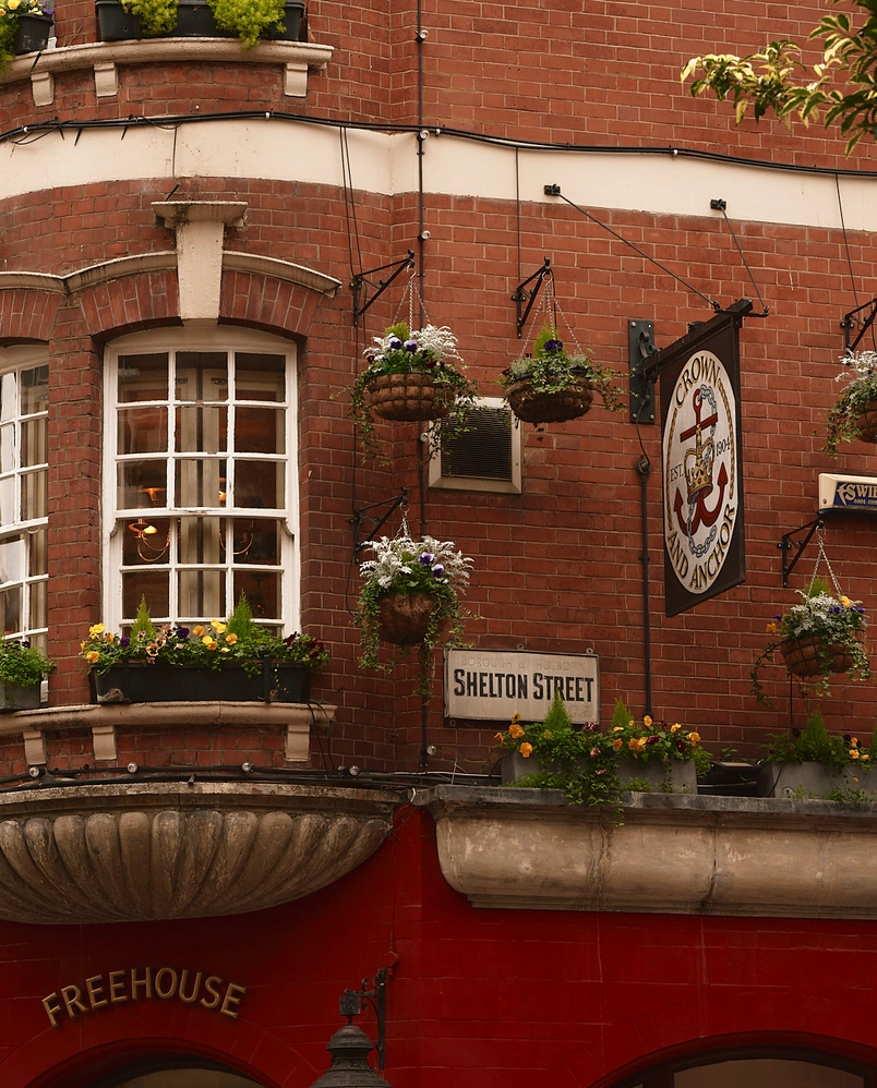 Red brick building corner with hanging flower baskets, window with flowers, and a Crown and Anchor pub sign on Shelton Street.