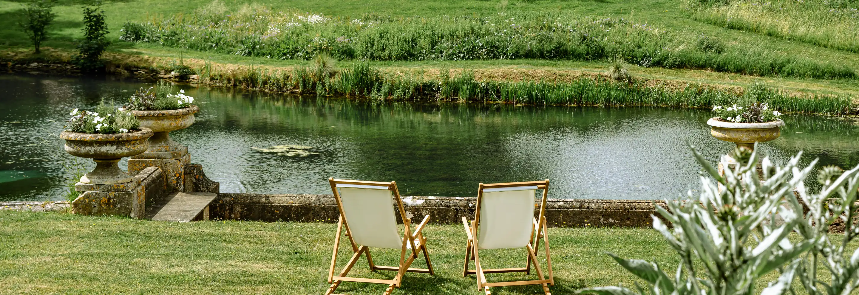 Two wooden deck chairs with white fabric facing a calm pond surrounded by lush green grass and plants.