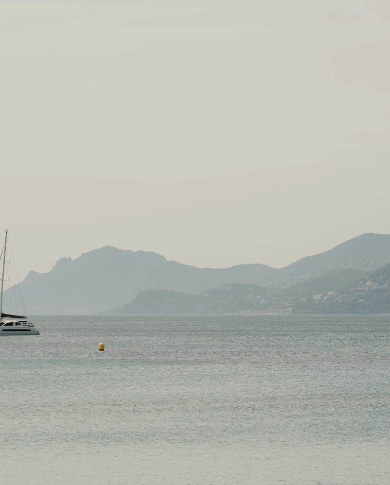 Sailboat on calm sea with mountainous coastline in the background under a cloudy sky.