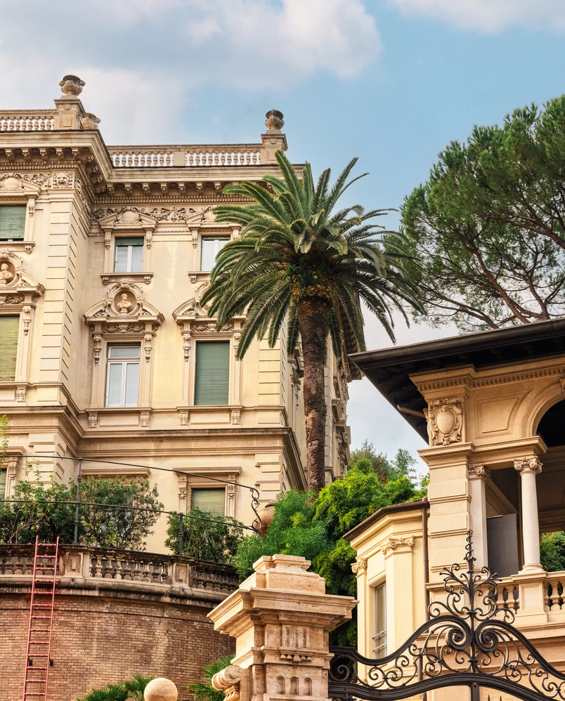 Ornate historic building with detailed architecture, palm tree, and wrought iron gate under blue sky with clouds.