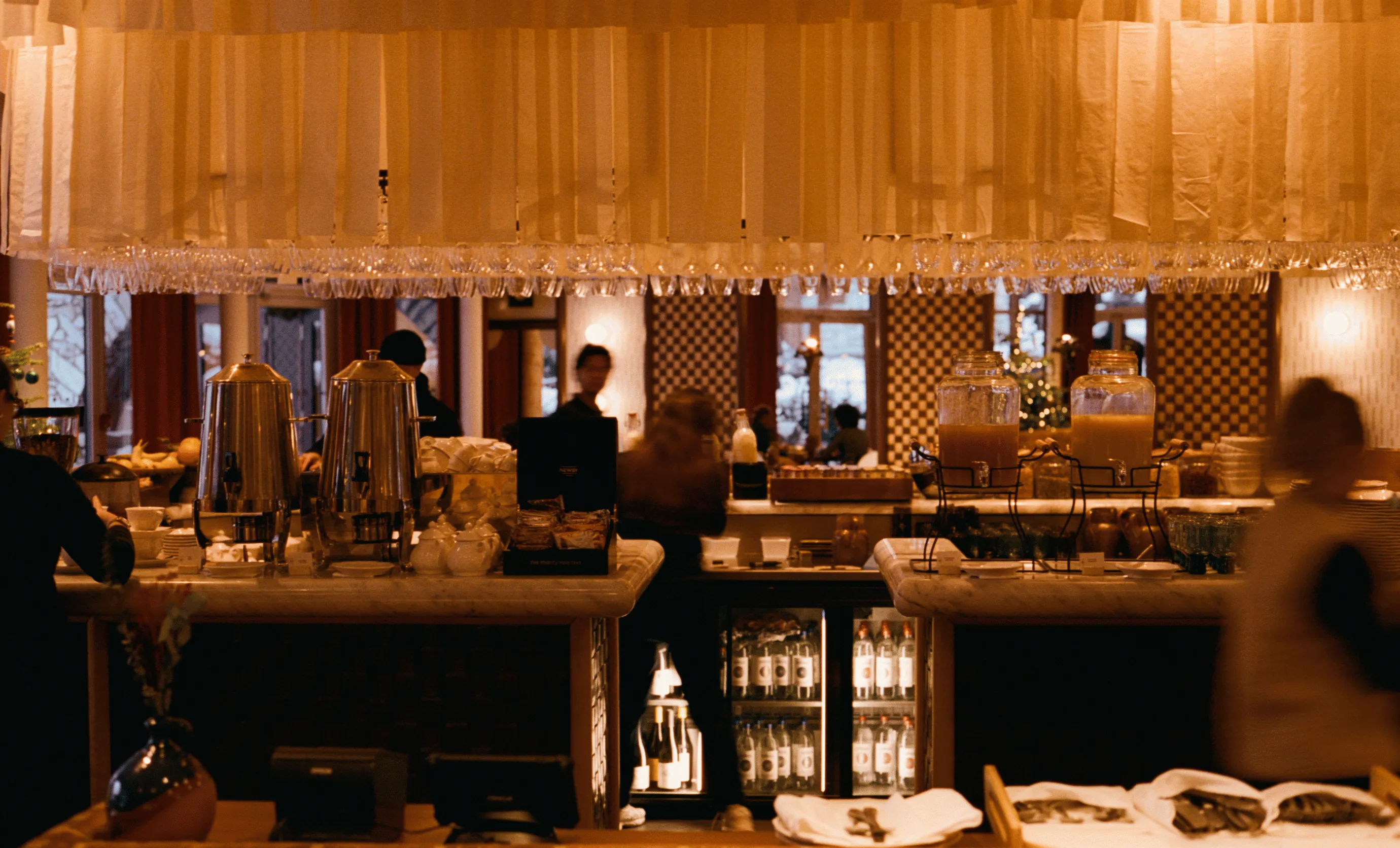 Cozy hotel bar with marble counters, beverage dispensers, and hanging glasses under warm lighting.