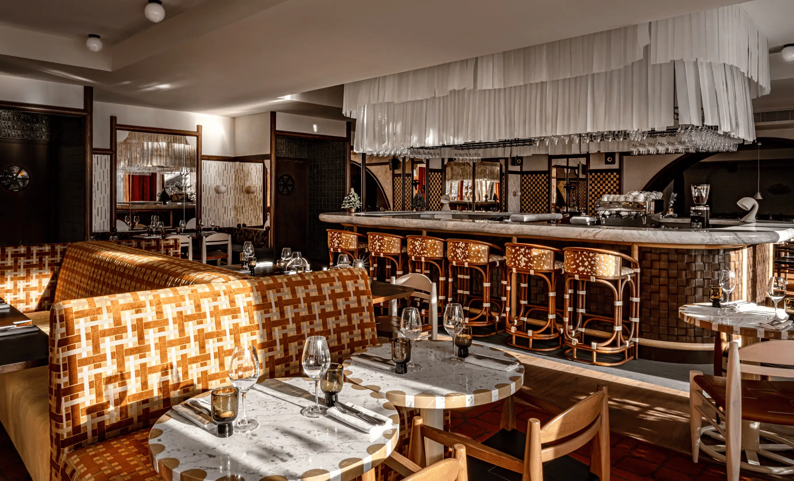 Elegant hotel bar area with marble-top tables, patterned orange cushioned seats, wooden chairs, and a backlit bar with hanging glassware.
