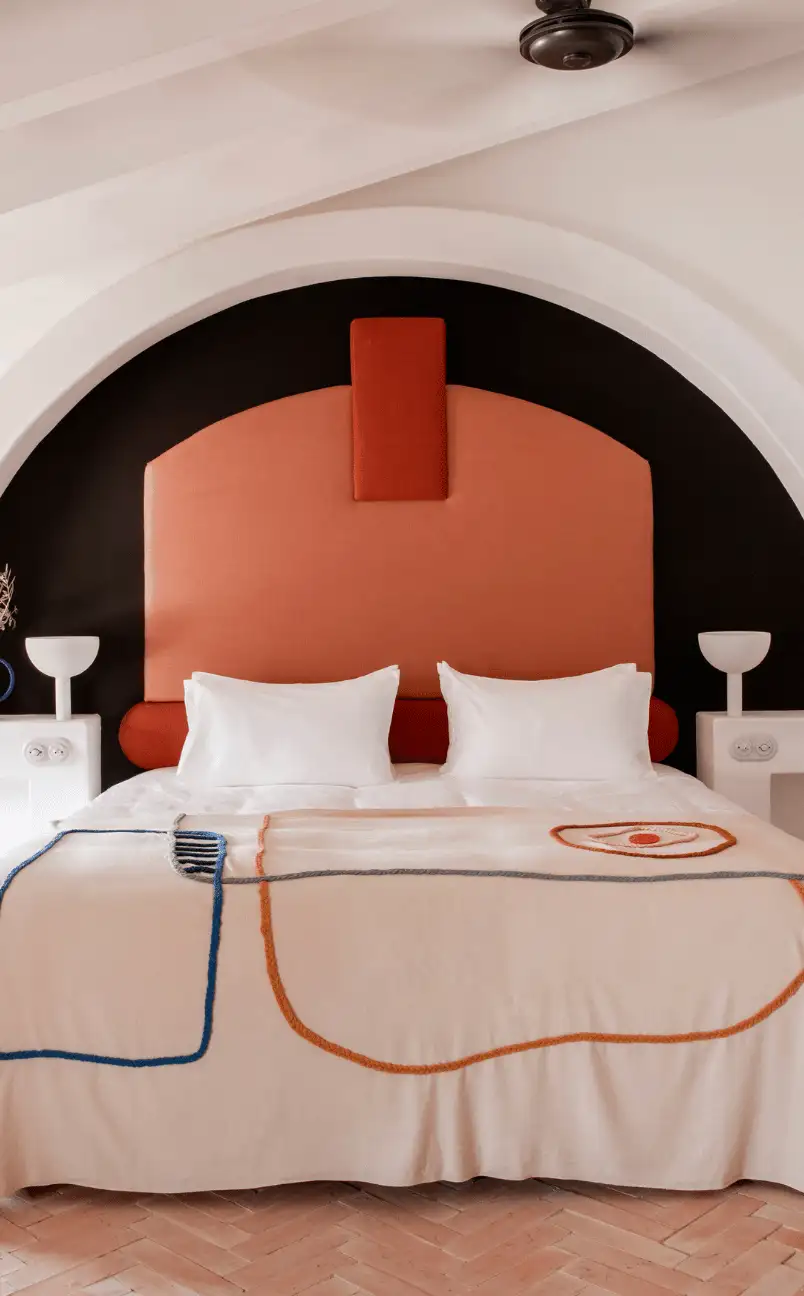 Modern hotel room with a large bed featuring a curved orange headboard and artistic bedding, flanked by white nightstands and lamps under an arch ceiling.