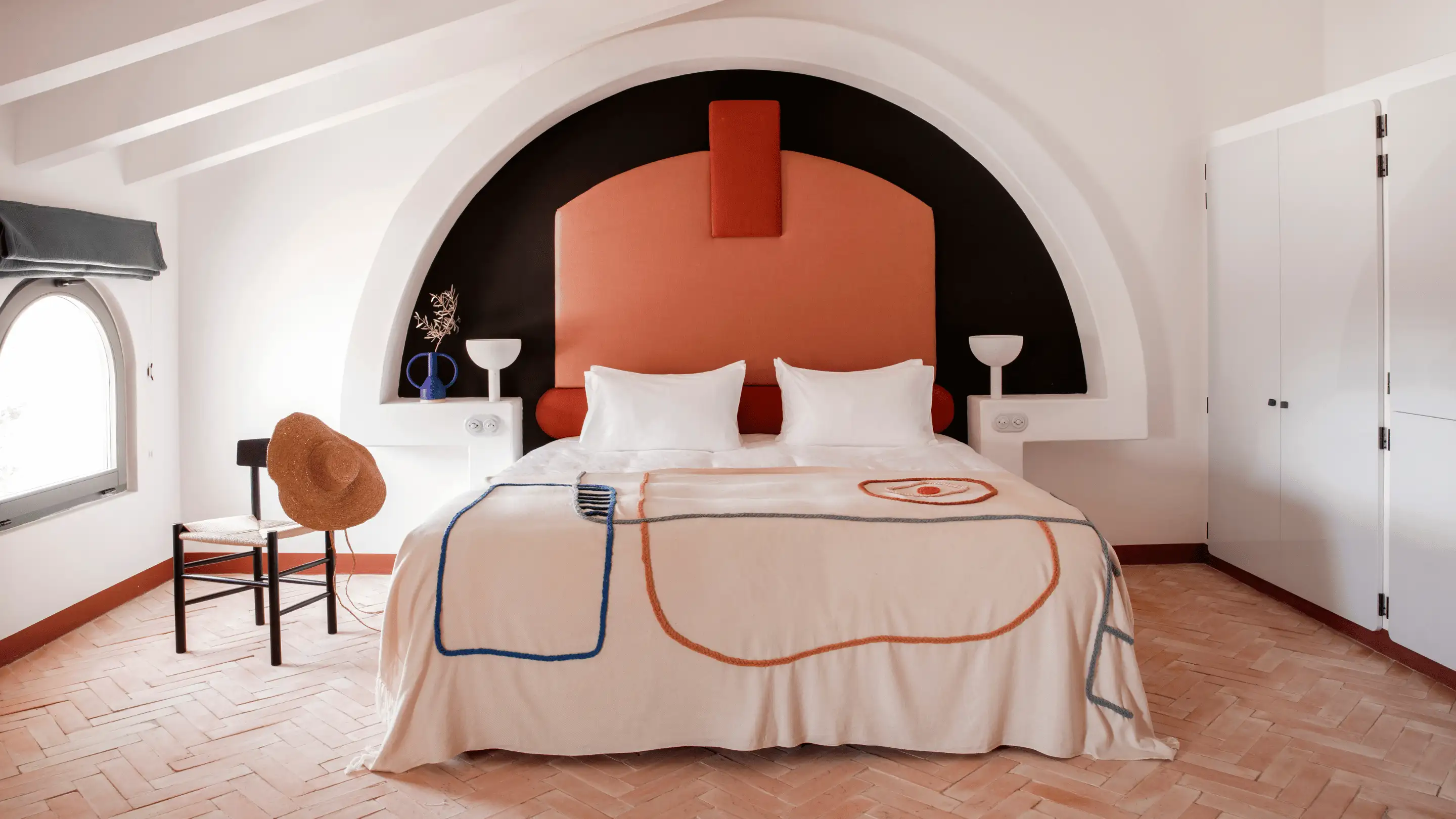 Minimalist bedroom with a large bed featuring a peach and black arched headboard, white pillows, and a patterned beige blanket, with a chair holding a straw hat next to an arched window.