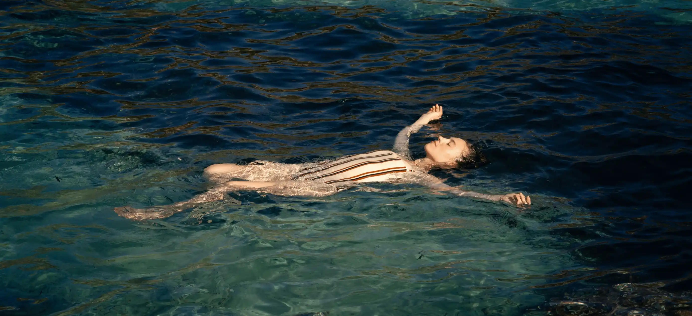 Woman floating relaxed on her back in clear blue water wearing a striped swimsuit.