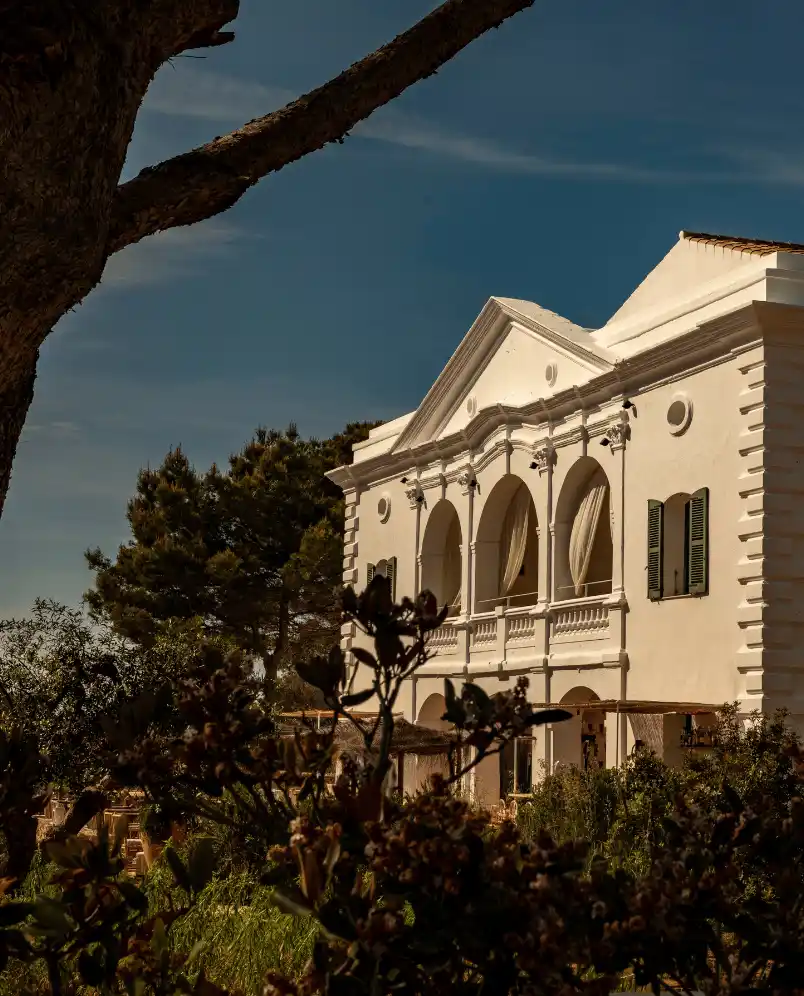 White historic-style building with arched windows and green shutters surrounded by lush trees and plants under a clear blue sky.