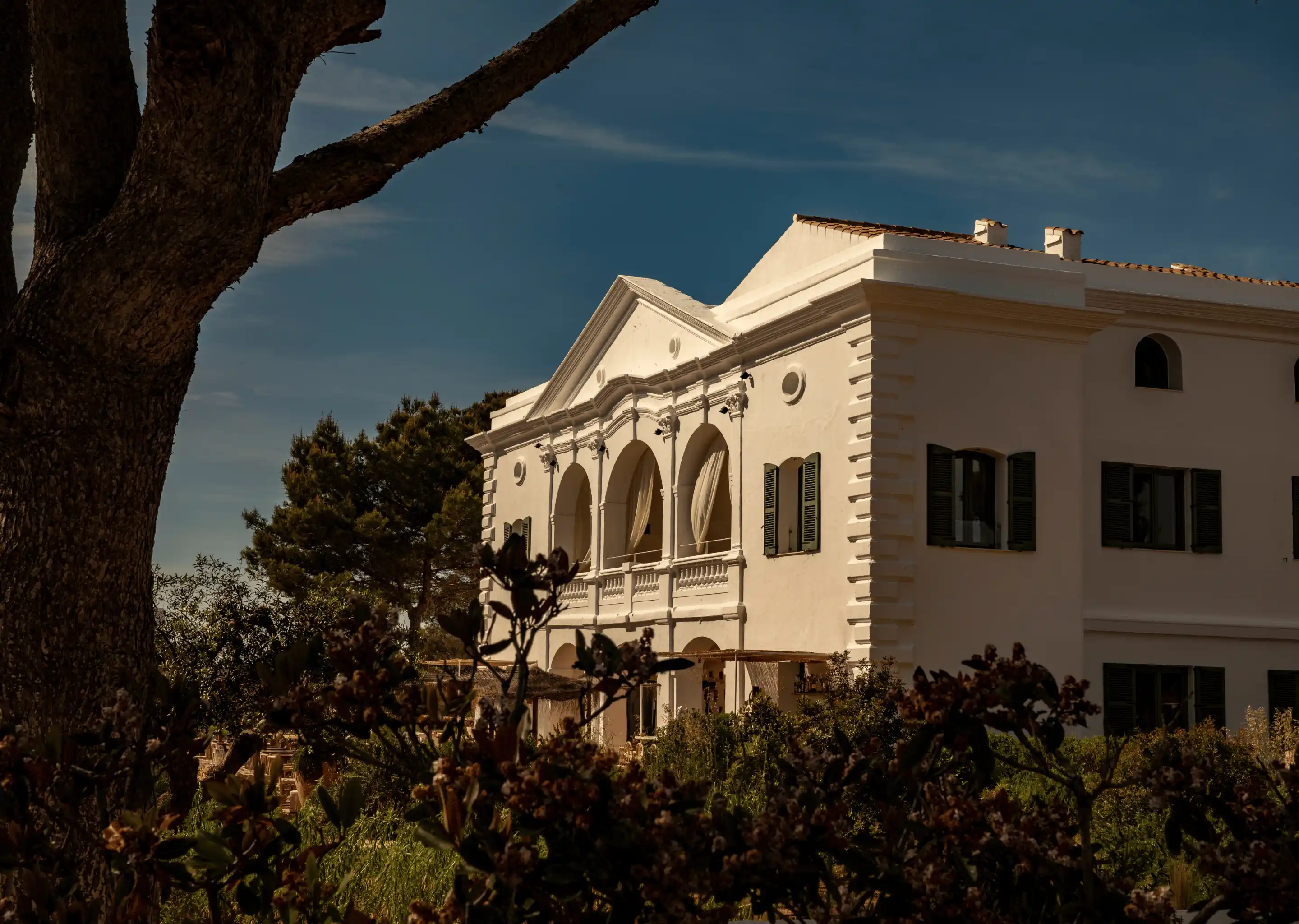 Elegant white building with arched balconies and green shutters surrounded by trees and bushes under a clear blue sky.