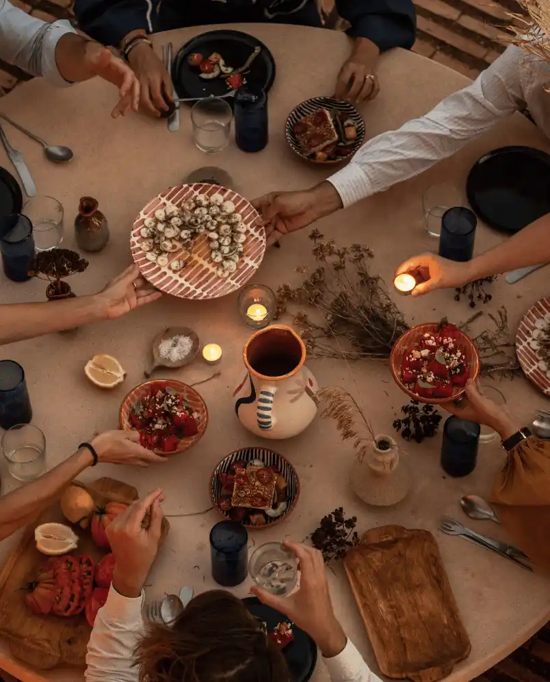 Overhead view of a group sharing food and lighting candles around a rustic table set with dishes, glasses, and dried flowers.