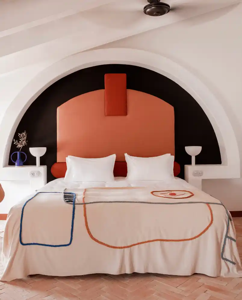 Modern bedroom with a large bed featuring white pillows and a cream blanket with abstract orange and blue designs, set against an arched wall with a black and terracotta color scheme.