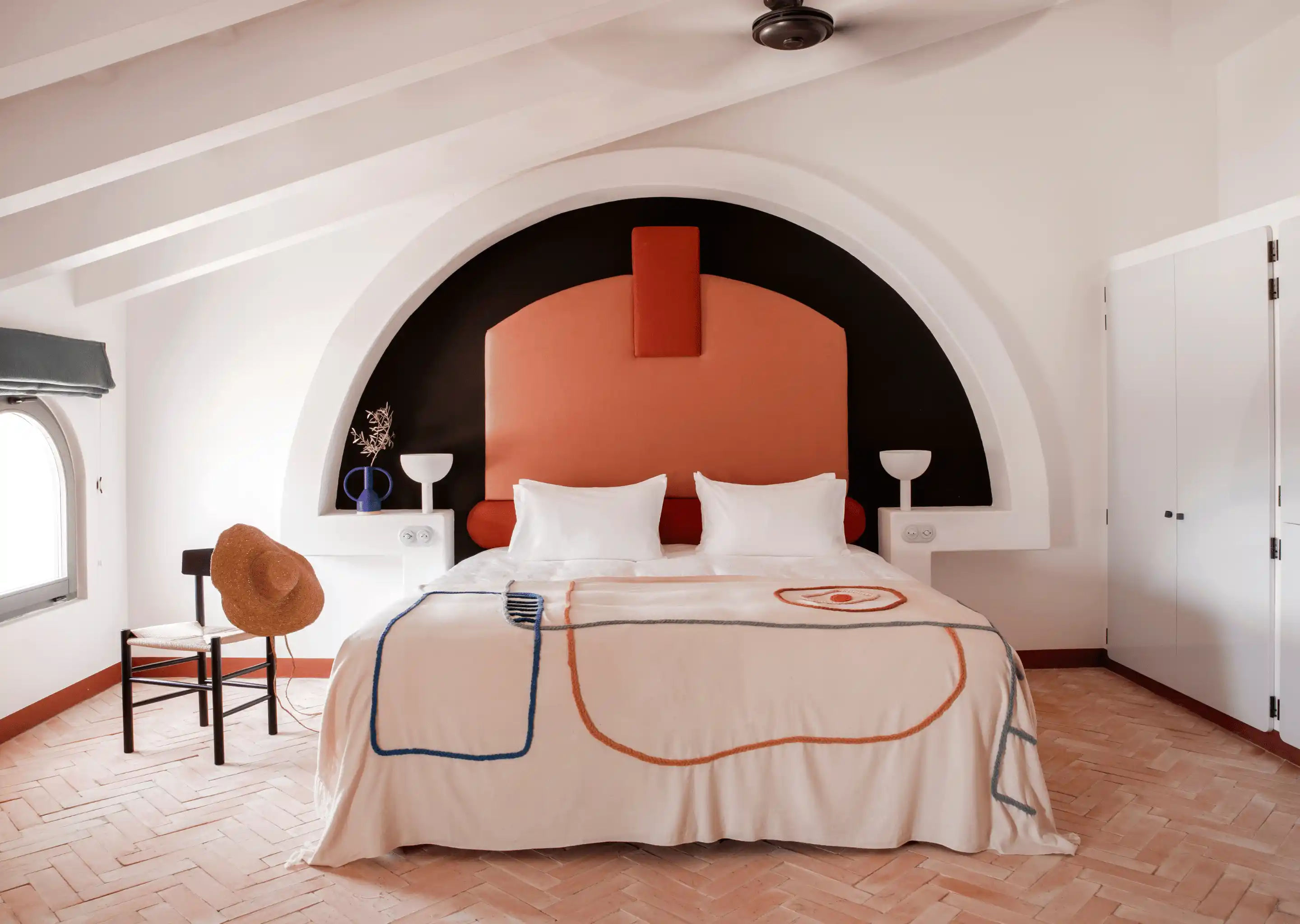 Minimalist hotel room with a double bed featuring an orange geometric headboard, patterned blanket, a chair with a straw hat, and white walls with an arched niche.