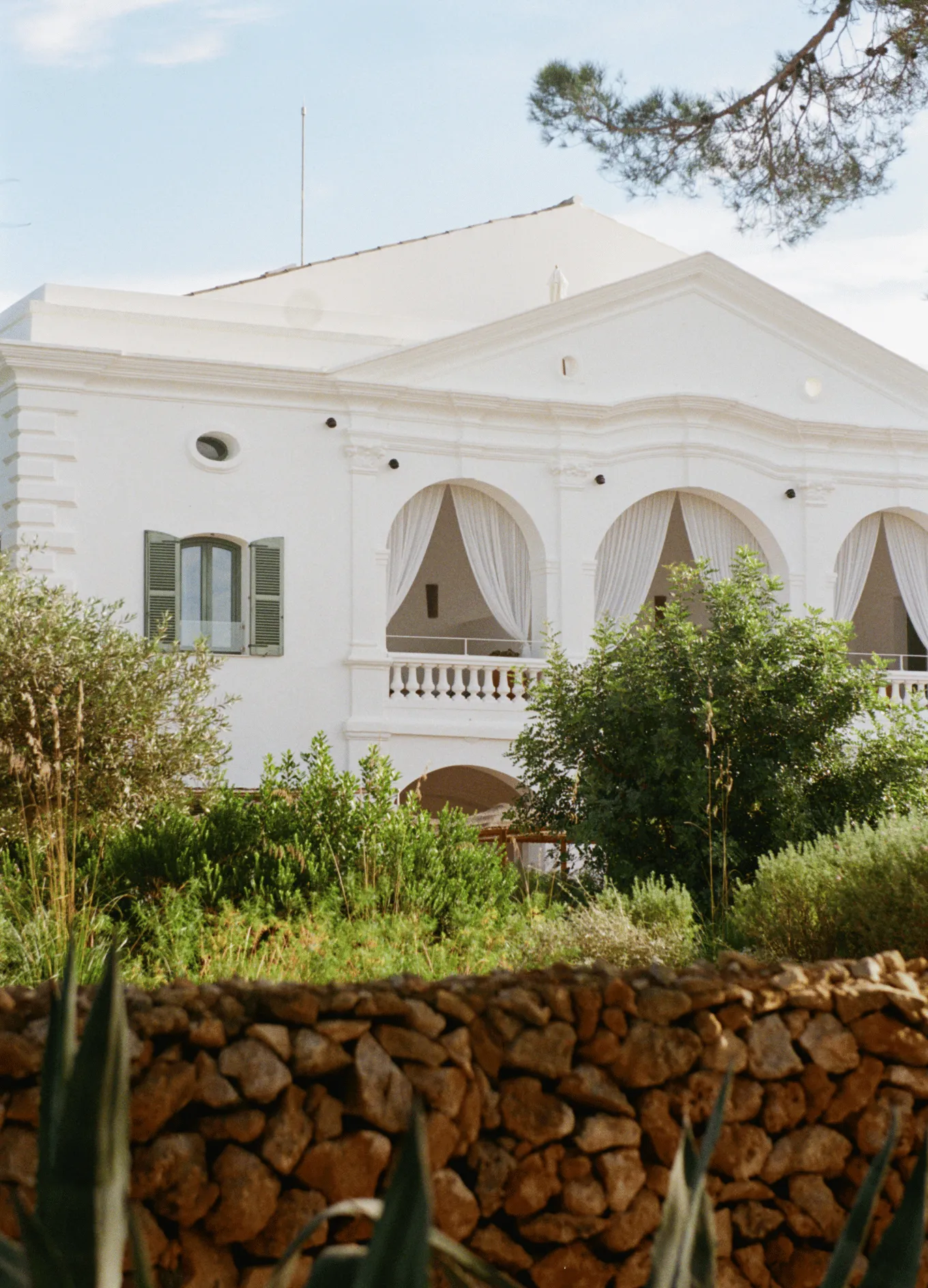White Mediterranean-style building with arches and balconies, surrounded by green shrubs and a stone wall in the foreground.