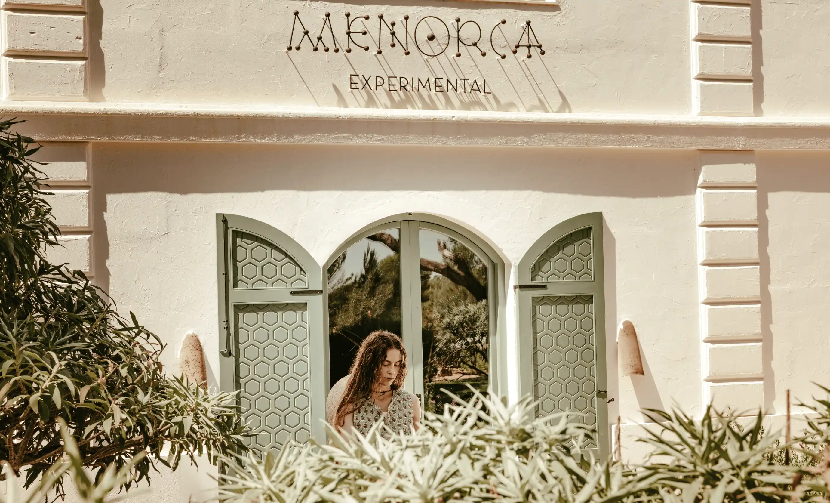 Woman with long hair standing behind green plants in front of a light-colored wall with arched green shutters and the words 'Menorca Experimental' above.