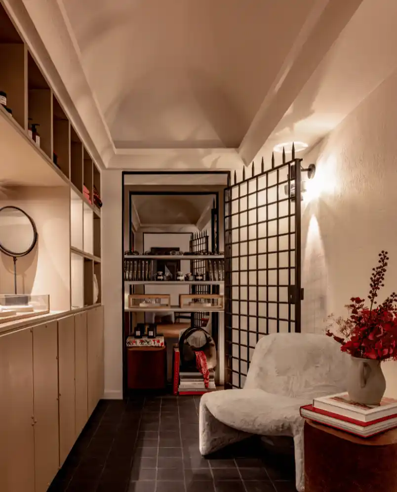 Cozy room with a soft white chair, a side table with stacked books and a vase of red flowers, wooden shelves, and a decorative black metal grid door.