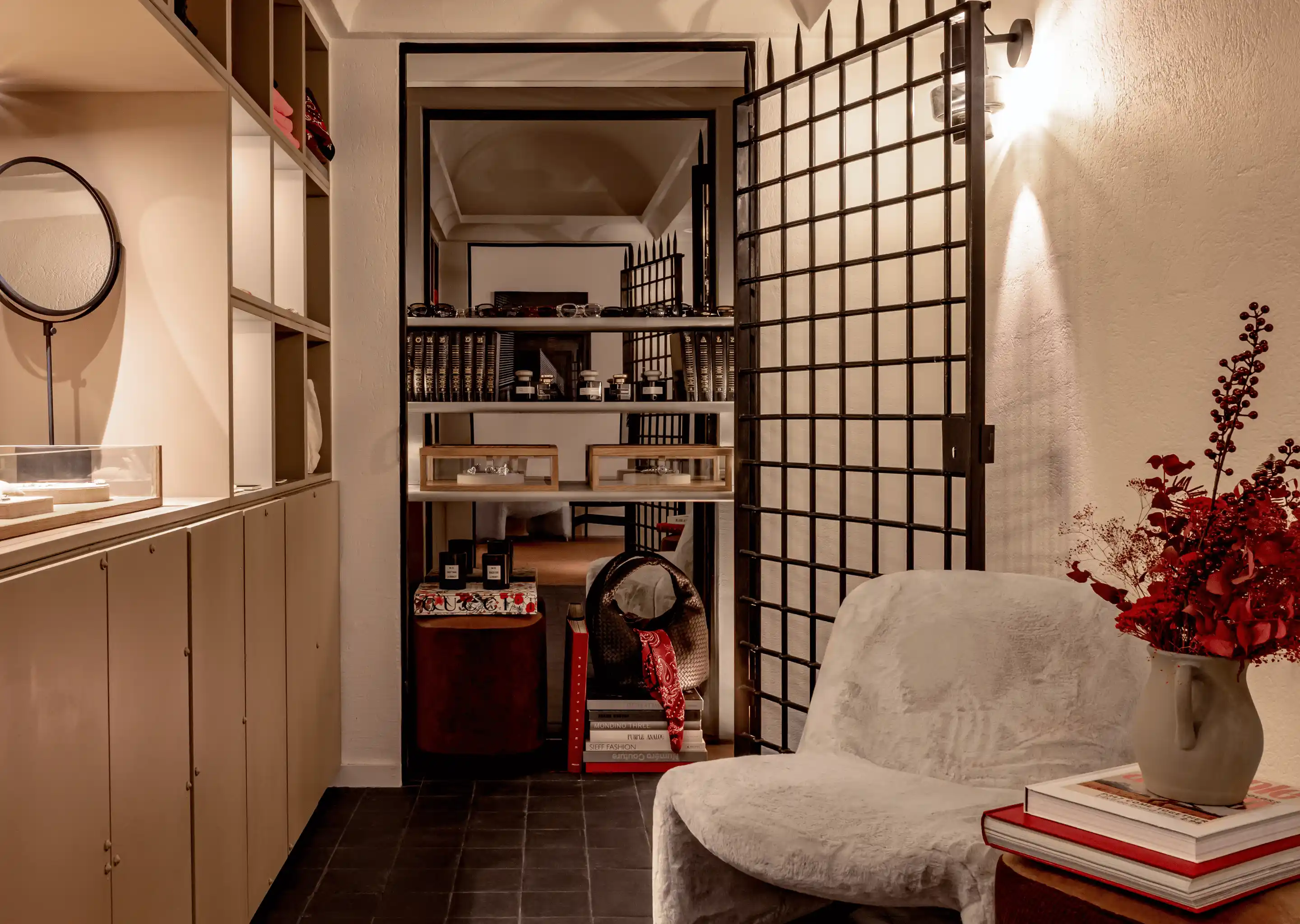 Cozy boutique room with a white covered armchair, a side table with stacked books and a vase with red flowers next to a black grid door and a shelf holding perfume bottles and glasses.