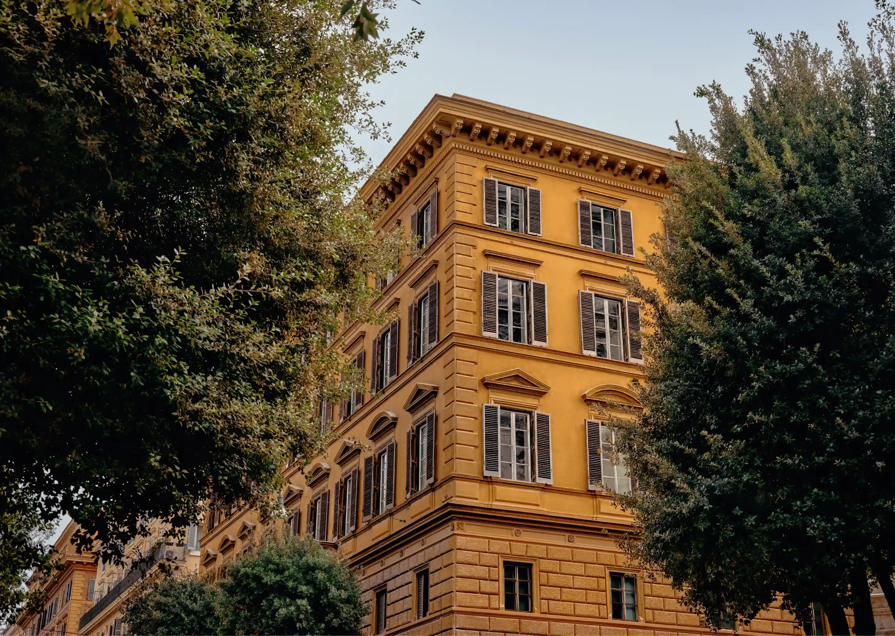 Yellow classical European building facade with multiple windows, partly framed by tall green trees under a clear sky.