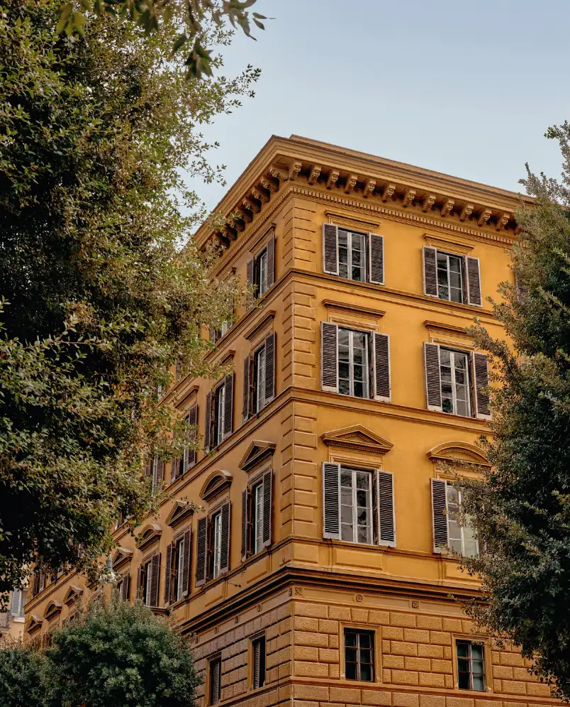 Corner view of a yellow ochre, multi-story historic building with black window shutters surrounded by green trees under a clear sky.