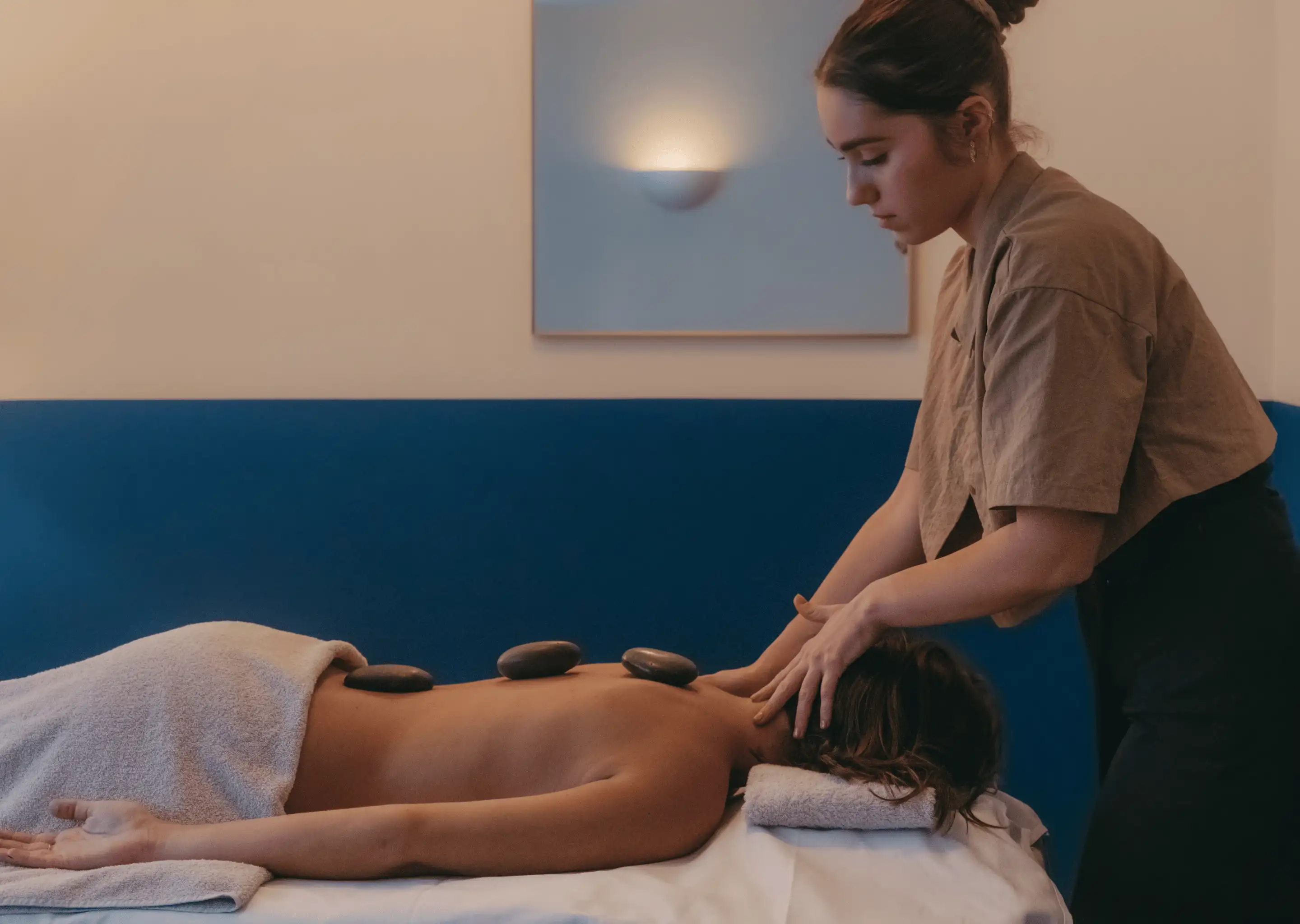Woman receiving a hot stone massage while lying face down with three stones placed on her back and therapist massaging her neck.