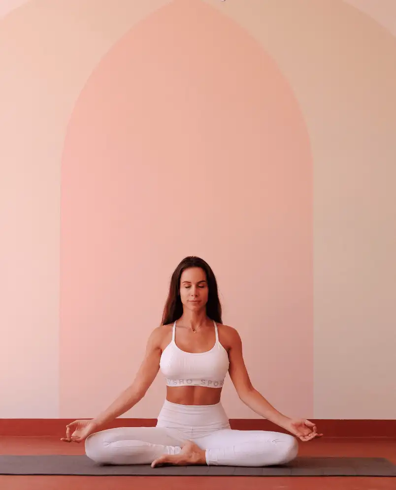 Woman in white sportswear meditating in a cross-legged yoga pose on a mat indoors with a soft pink arched background.