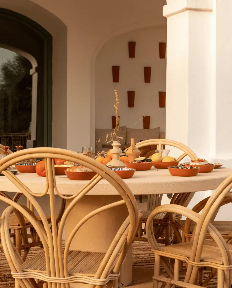 Outdoor dining setup with a round stone table surrounded by rattan chairs, decorated with clay bowls and vases on a warm-toned patio.