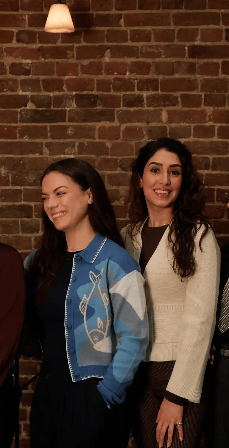 Two women smiling and standing side by side in front of a brick wall with a hanging light above.