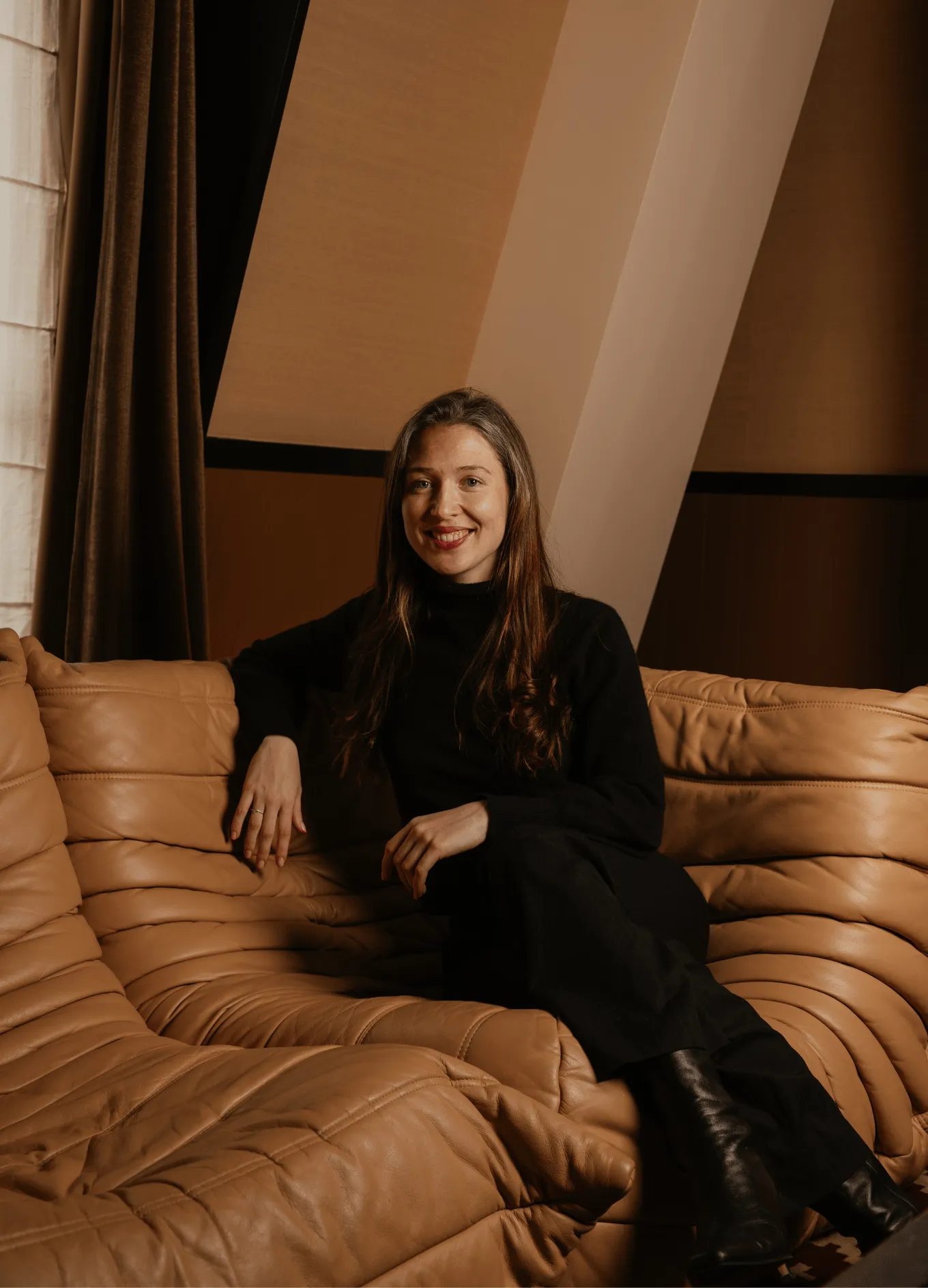 Smiling woman with long brown hair wearing black sitting on a tan leather couch in a warmly lit room.