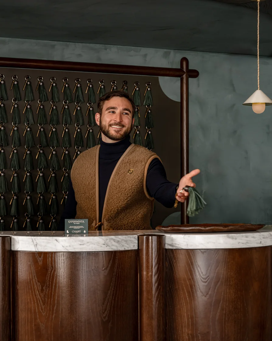 Smiling man in brown vest stands behind marble and wood counter holding out a key with green tassel in a modern interior.