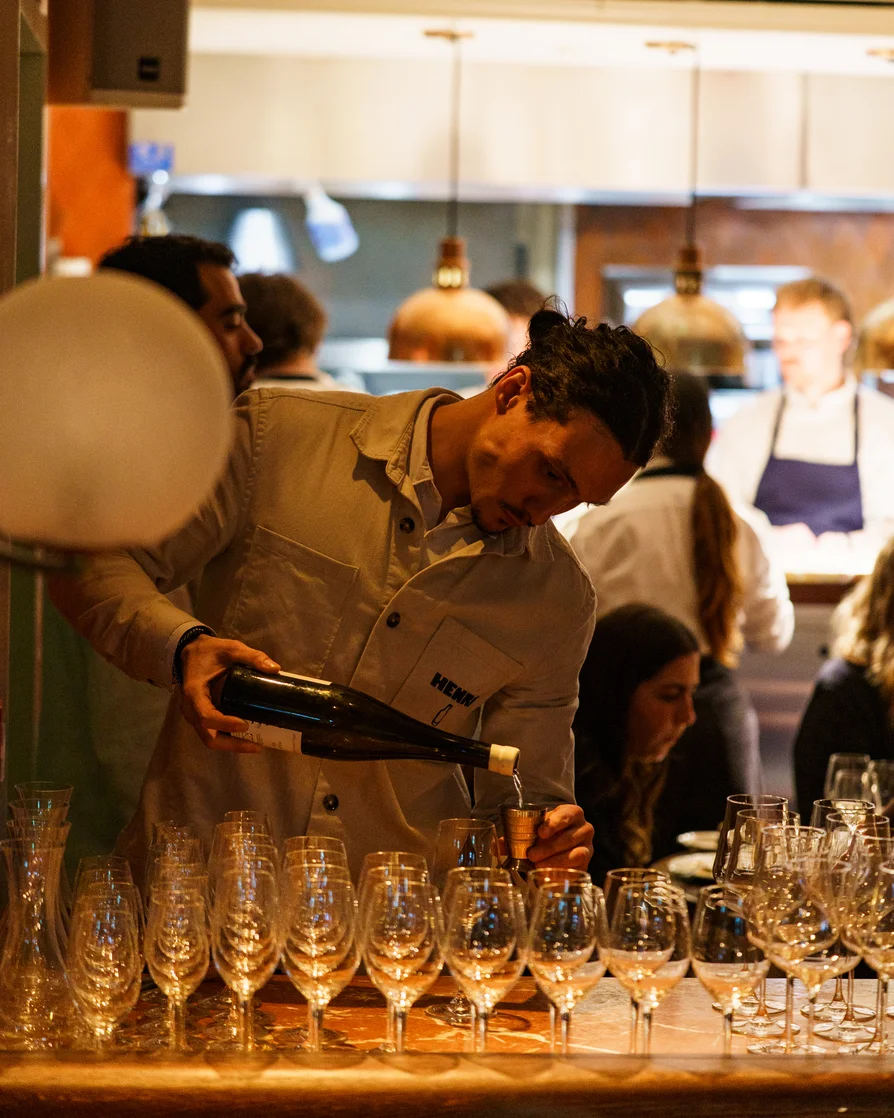 Bartender pouring wine into a jigger behind a bar with rows of empty wine glasses in front.