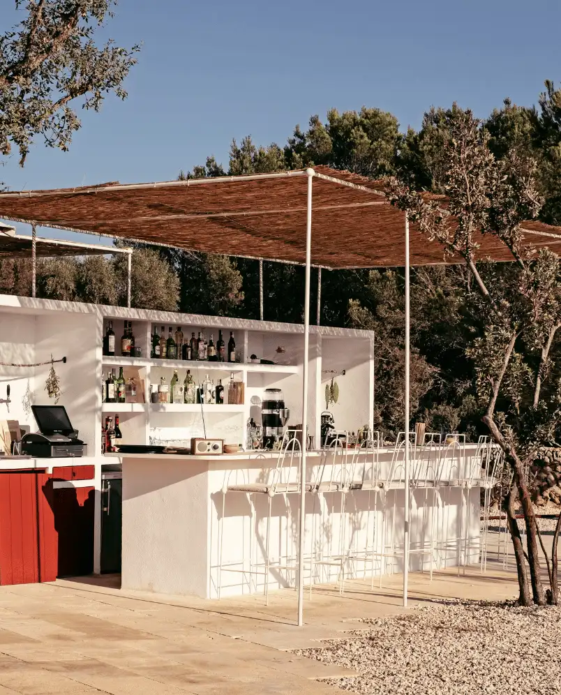 Outdoor bar with white counter, several bottles on shelves, and metal bar stools under a woven canopy surrounded by trees.