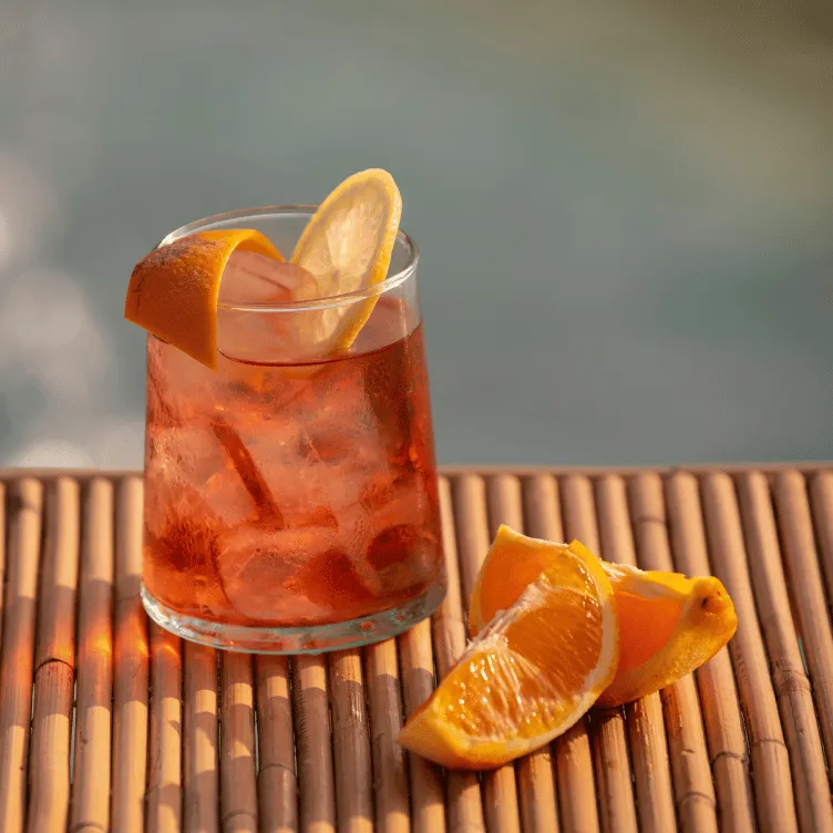 Glass of iced orange cocktail garnished with a lemon slice on a bamboo surface next to orange wedges.